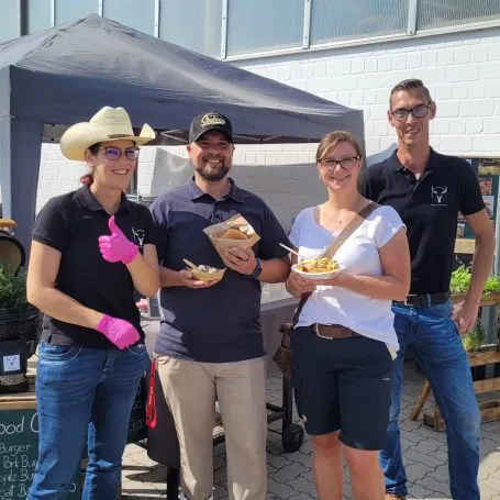 Vier Personen mit frisch zubereiteten Speisen am Wild Black BBQ Stand unter dem Pavillon