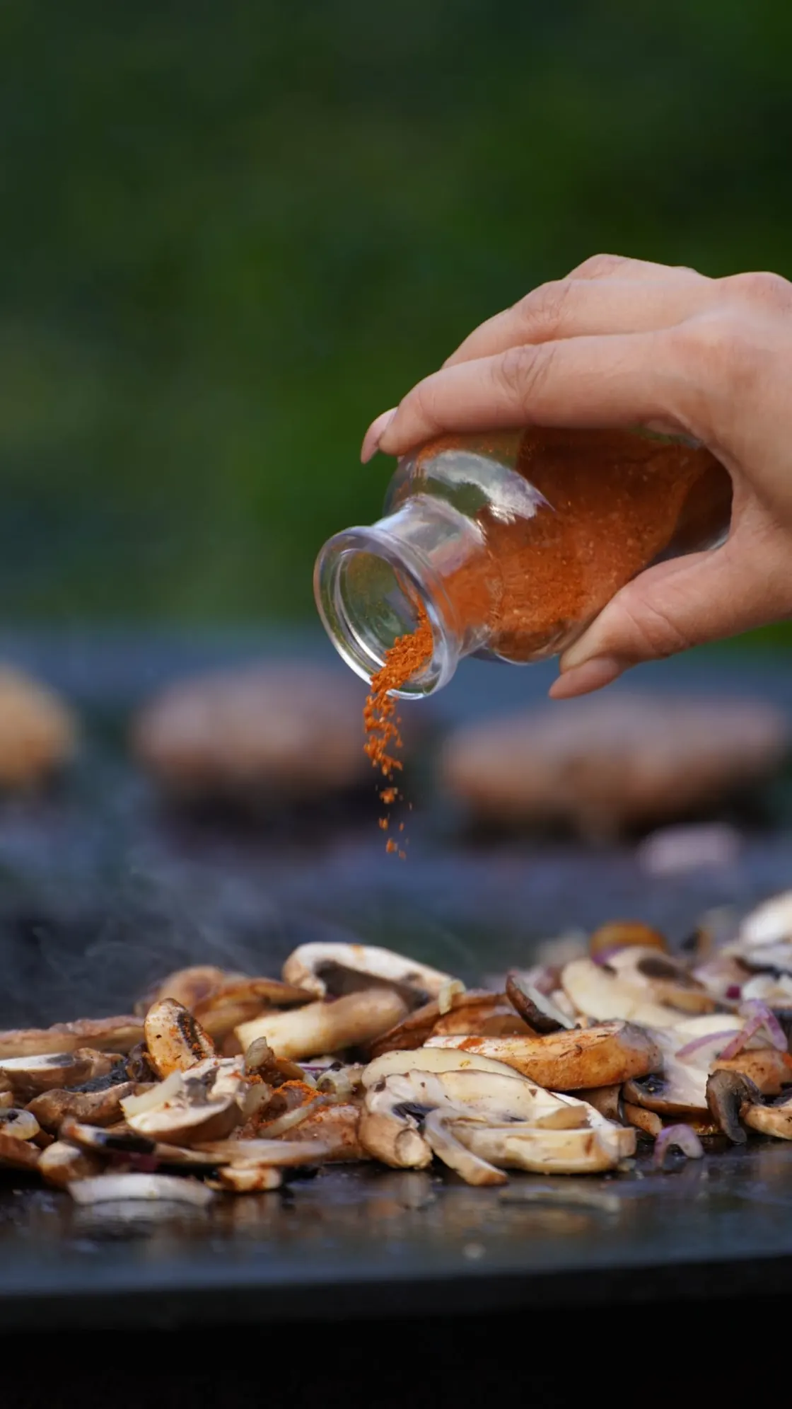 Gewürz wird aus einem Glas über brutzelnde Champignons auf der Plancha gestreut