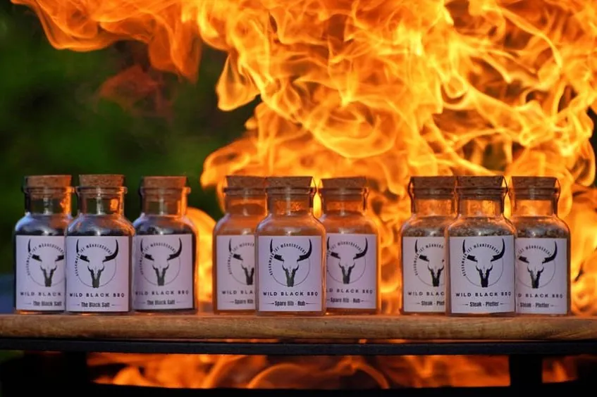 Reihe eigener Gewürzgläser von Wild Black BBQ vor lodernden Flammen